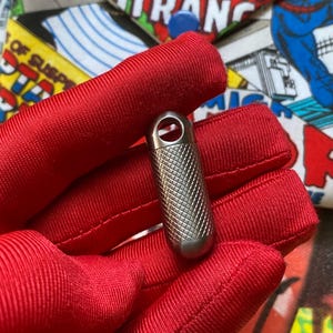 May include: A small, silver-colored metal capsule with a textured, diamond-patterned surface. The capsule has a rounded bottom and a loop at the top. The capsule is held in a red fabric. Comic book pages are in the background.