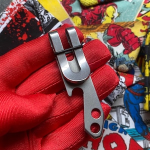 May include: A silver pocket clip with a bottle opener and two circular holes. The clip is held by a red glove against a backdrop of comic book panels featuring colorful illustrations of superheroes.