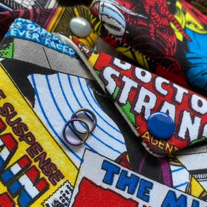 May include: Close-up of comic book fabric items with button closures. The fabric features colorful comic book panels with text like "Suspense" and "Doctor Strange." Two small, iridescent rings are in the foreground.