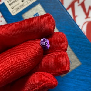 May include: A small, purple metal object with a circular design. The object is held in a red fabric. The background includes a blue surface and a white and red design.
