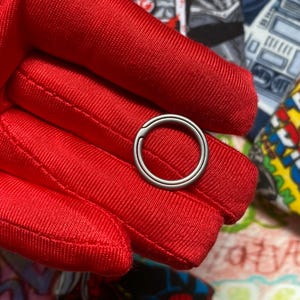May include: A silver-colored, circular ring with a hinged opening. The ring is held against a bright red fabric, with a patterned background visible. The ring appears to be made of metal and has a smooth, polished surface.