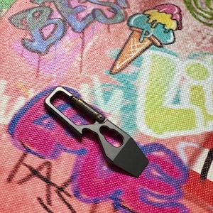 May include: A gray and silver multi-tool with a bottle opener and a small blade. The tool has a spring-loaded mechanism and a rectangular blade. The background features graffiti art with an ice cream cone.