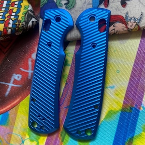 May include: Two bright blue metal knife handle scales featuring a diagonal ribbed texture. Each piece includes pre-drilled circular holes for assembly, with a curved ergonomic shape designed for a folding knife handle replacement set.
