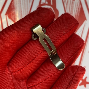 May include: A metallic pocket clip with a brushed brass finish, featuring a curved design, a central elongated slot, and two small circular mounting holes at the top end, presented against a bright red fabric background.