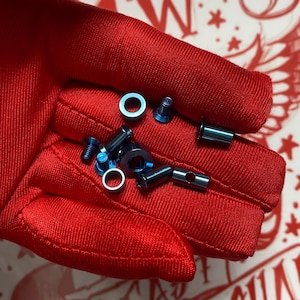 May include: A collection of small, metallic blue hardware components including several cylindrical spacers, flat-head screws, and circular washers. These industrial fasteners feature a smooth, iridescent finish and are arranged in a cluster.