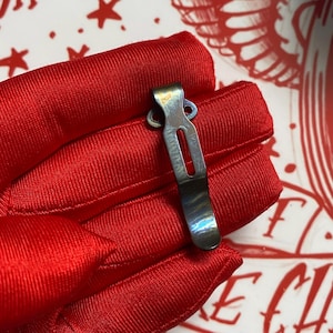 May include: A small, metallic pocket clip with a rectangular slot and two circular holes. The clip has a gradient finish, appearing silver and blue. It rests on a vibrant red, textured fabric, creating a striking contrast.