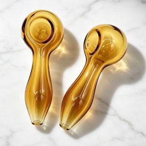 May include: Two amber-colored glass pipes with a rounded bowl and elongated stem. The pipes are made of transparent glass, allowing light to pass through. They are placed on a white marble surface.