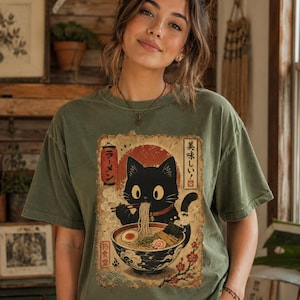 May include: Olive green t-shirt featuring a black cat enjoying ramen. The graphic includes Japanese text and a red sun design. The cat is depicted with large eyes and is holding chopsticks. The shirt has a vintage, worn look.