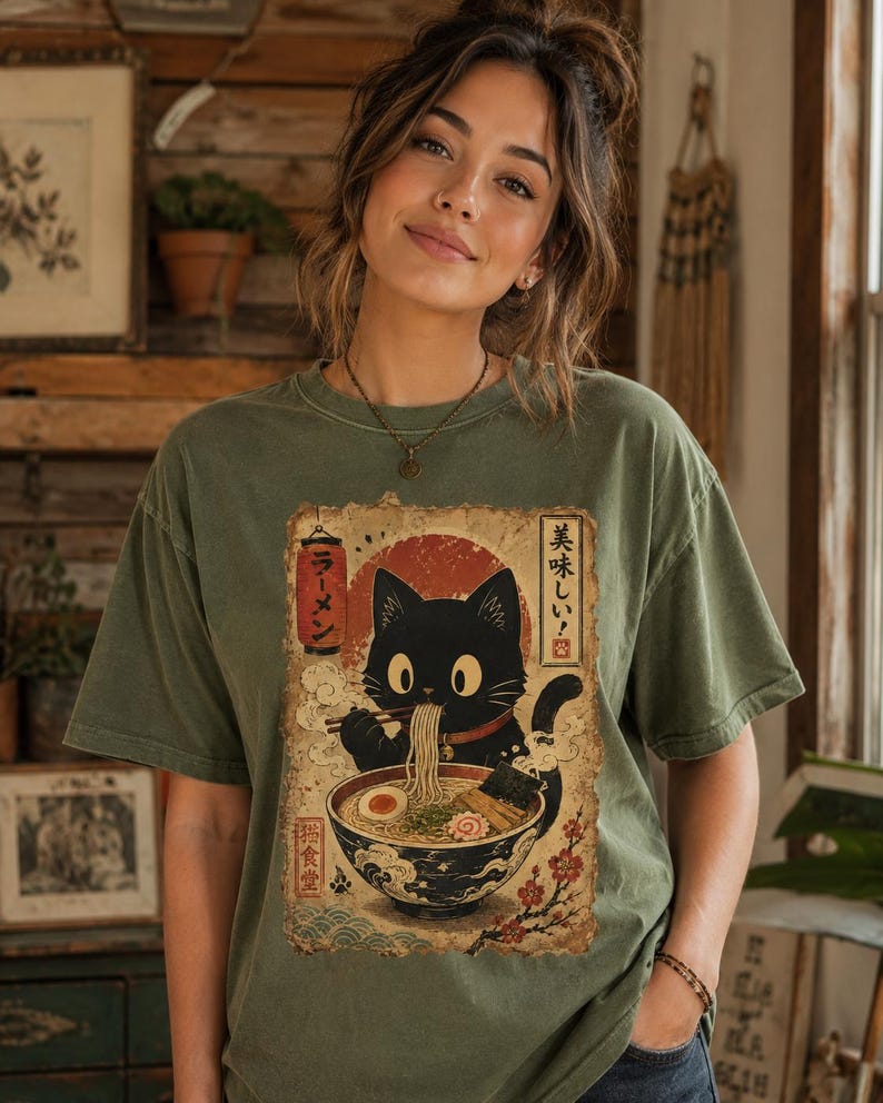 May include: Olive green t-shirt featuring a black cat enjoying ramen. The graphic includes Japanese text and a red sun design. The cat is depicted with large eyes and is holding chopsticks. The shirt has a vintage, worn look.
