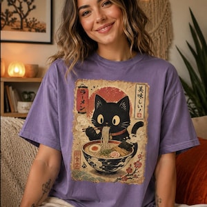 May include: A purple t-shirt featuring a vintage-style illustration of a black cat enjoying ramen. The cat is depicted with large eyes and is holding chopsticks. Japanese text is visible in the design. The shirt has short sleeves.