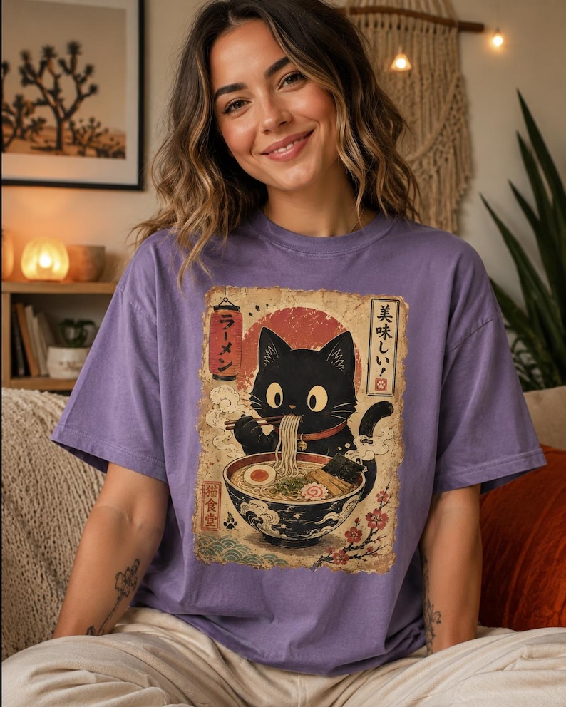 May include: A purple t-shirt featuring a vintage-style illustration of a black cat enjoying ramen. The cat is depicted with large eyes and is holding chopsticks. Japanese text is visible in the design. The shirt has short sleeves.