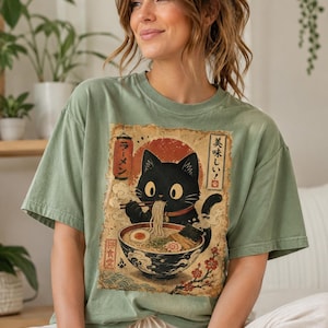 May include: Sage green t-shirt featuring a vintage-style graphic of a black cat enjoying ramen. The graphic includes Japanese text and a red sun. The cat is depicted with a red collar, and the ramen bowl is filled with noodles, egg, and seaweed.