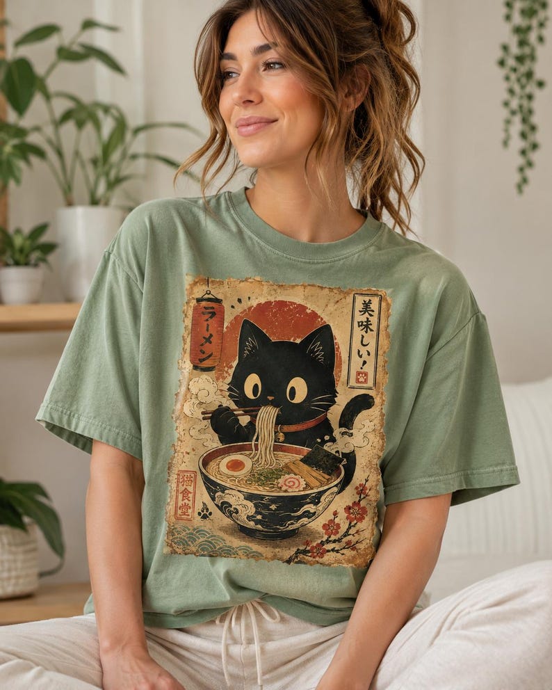 May include: Sage green t-shirt featuring a vintage-style graphic of a black cat enjoying ramen. The graphic includes Japanese text and a red sun. The cat is depicted with a red collar, and the ramen bowl is filled with noodles, egg, and seaweed.