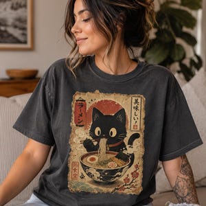 May include: Dark gray t-shirt featuring a black cat enjoying ramen. The graphic includes Japanese text and a red sun design. The cat is depicted with large eyes and is holding chopsticks. The shirt is a casual style.