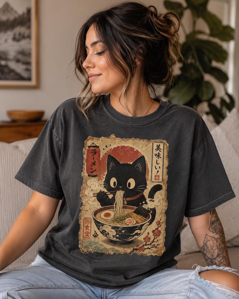 May include: Dark gray t-shirt featuring a black cat enjoying ramen. The graphic includes Japanese text and a red sun design. The cat is depicted with large eyes and is holding chopsticks. The shirt is a casual style.