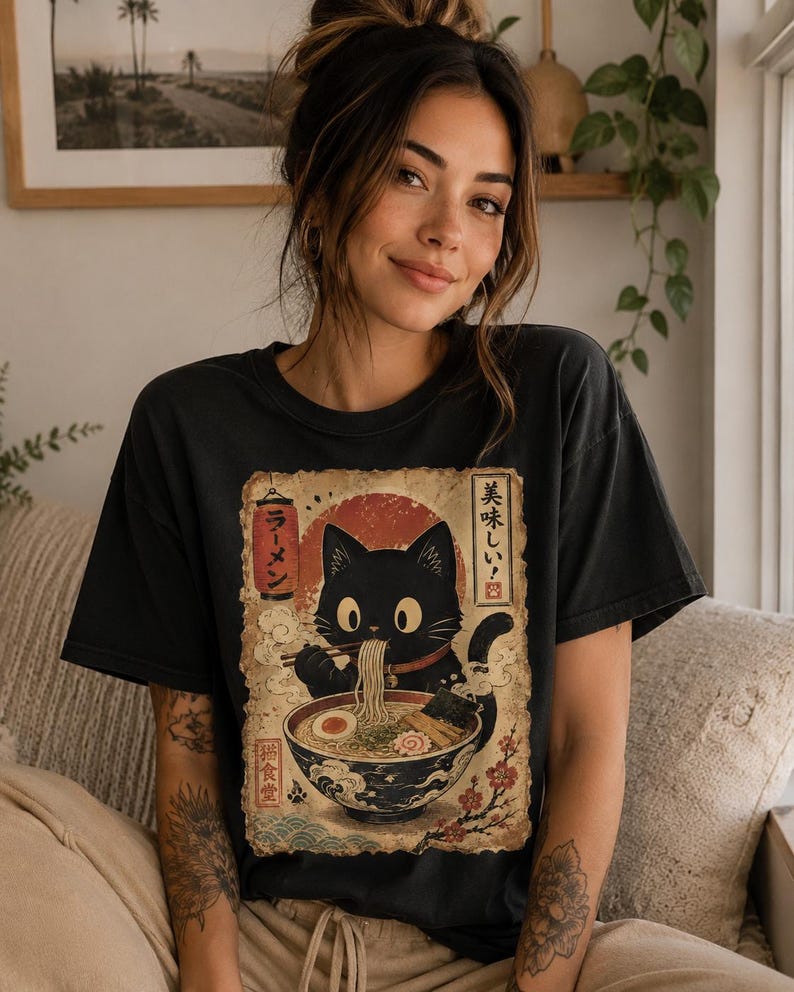 May include: Black t-shirt featuring a vintage-style illustration of a black cat eating ramen. The graphic includes Japanese text and a red sun. The shirt is a casual, comfortable style.