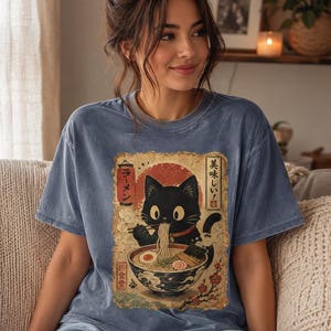 May include: Blue t-shirt featuring a black cat enjoying ramen from a bowl. The graphic has Japanese text and a red sun design. The cat is holding chopsticks with noodles. The shirt is a comfortable, casual style.