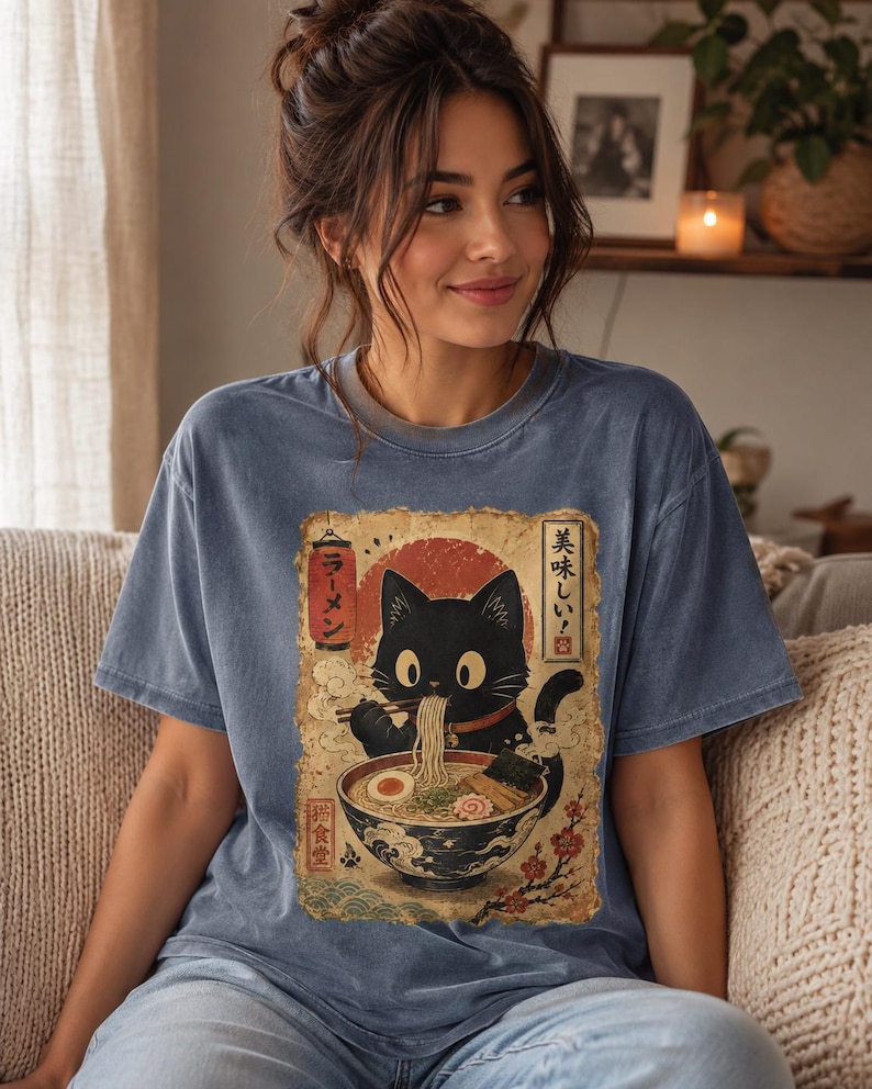 May include: Blue t-shirt featuring a black cat enjoying ramen from a bowl. The graphic has Japanese text and a red sun design. The cat is holding chopsticks with noodles. The shirt is a comfortable, casual style.