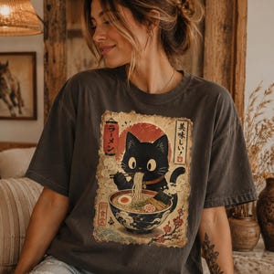 May include: Dark gray t-shirt featuring a black cat eating ramen from a bowl. The graphic includes Japanese text and a red sun design. The shirt has a vintage, worn look.