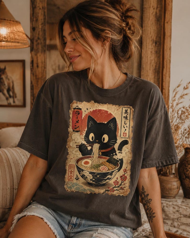 May include: Dark gray t-shirt featuring a black cat eating ramen from a bowl. The graphic includes Japanese text and a red sun design. The shirt has a vintage, worn look.