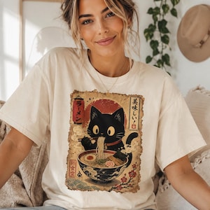 May include: Cream-colored t-shirt featuring a black cat enjoying ramen from a bowl. The graphic has Japanese text and a red sun design. The cat is eating noodles with chopsticks. The shirt is a casual, comfortable style.