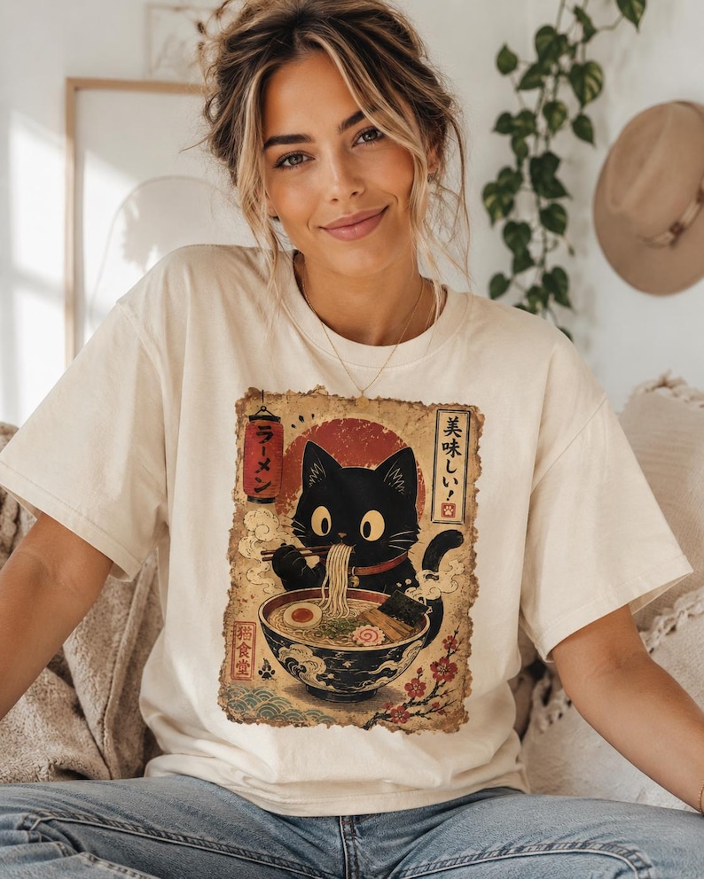 May include: Cream-colored t-shirt featuring a black cat enjoying ramen from a bowl. The graphic has Japanese text and a red sun design. The cat is eating noodles with chopsticks. The shirt is a casual, comfortable style.
