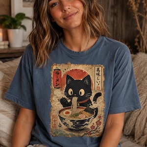 May include: Blue t-shirt featuring a black cat enjoying ramen. The graphic has Japanese text and a red sun design. The cat is depicted with large eyes and is holding chopsticks. The shirt is a relaxed fit.