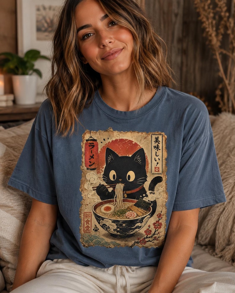 May include: Blue t-shirt featuring a black cat enjoying ramen. The graphic has Japanese text and a red sun design. The cat is depicted with large eyes and is holding chopsticks. The shirt is a relaxed fit.