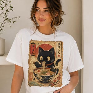 May include: White t-shirt featuring a black cat enjoying ramen from a bowl. The graphic includes Japanese text and a red sun design. The cat is depicted with large eyes and is holding chopsticks. The shirt is a casual style.