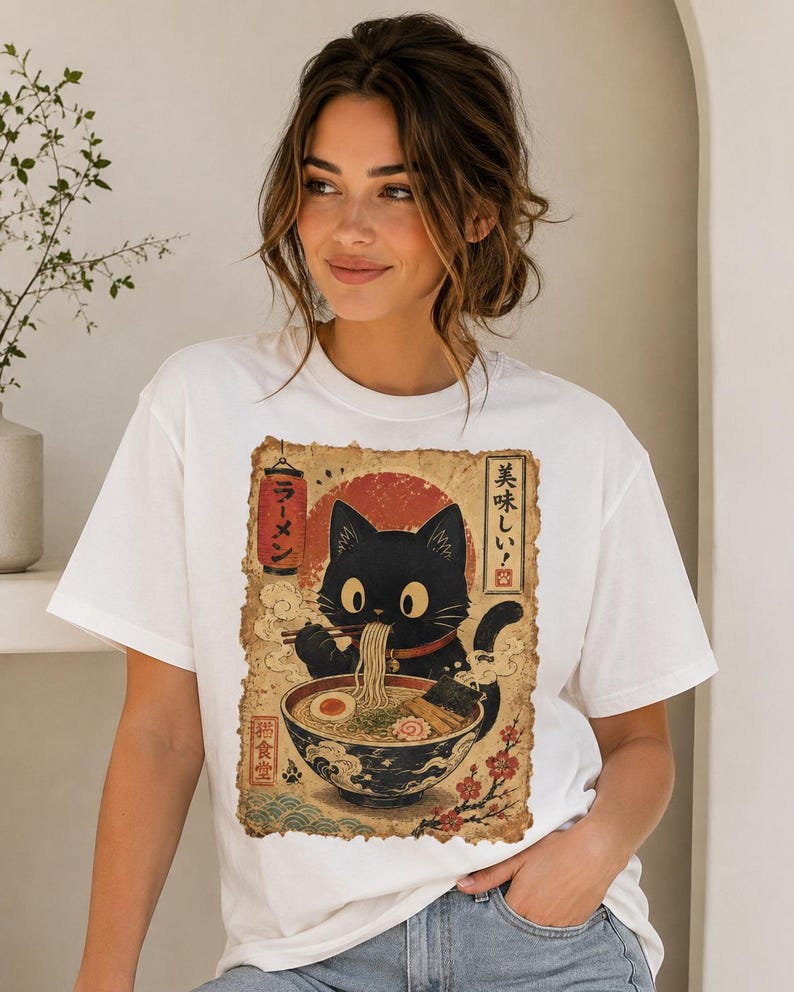 May include: White t-shirt featuring a black cat enjoying ramen from a bowl. The graphic includes Japanese text and a red sun design. The cat is depicted with large eyes and is holding chopsticks. The shirt is a casual style.