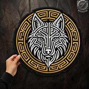 May include: A circular embroidered patch featuring a stylised white wolf head with geometric lines centred within a gold and black Greek key border. The design is stitched with high-contrast thread on a black fabric base, creating a detailed emblem.