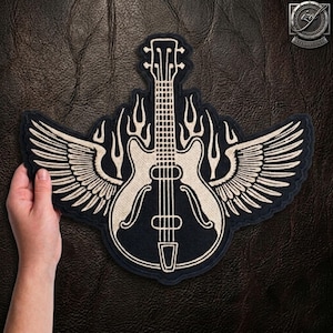 Puede incluir: Un parche bordado en negro y blanco roto que presenta un diseño central de guitarra eléctrica flanqueado por dos alas grandes y detalladas y motivos de llamas estilizados que surgen del cuerpo de la guitarra, todo con un borde oscuro sobre fondo texturizado.