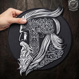 Pode incluir: Um emblema bordado circular apresentando o perfil prateado de um guerreiro barbudo com cabelos longos e fluidos, usando um capacete ornamentado com padrões geométricos intrincados e volutas sobre um fundo de tecido preto.