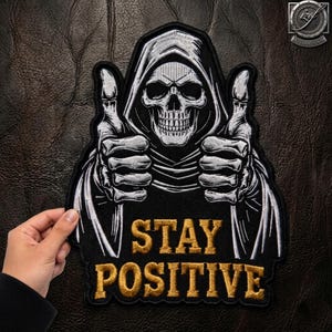 Könnte beinhalten: Ein schwarz-weiß bestickter Aufnäher mit einem Skelett in einer Kapuze, das beide Hände mit erhobenen Daumen zeigt. Unter dem Skelett ist der Text STAY POSITIVE in goldenem Garn auf schwarzem Hintergrund gestickt.