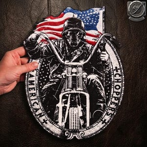 Puede incluir: Un parche bordado en blanco y negro que muestra una calavera con casco montada en una motocicleta. El diseño incluye una bandera estadounidense ondeando al fondo y el texto AMERICAN CHOPPERS impreso a lo largo del borde ovalado.