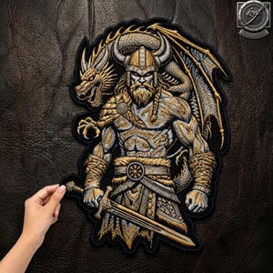 Pode incluir: Um grande emblema bordado apresentando um guerreiro barbudo usando um capacete com chifres e armadura com acabamento em pele, segurando uma espada. Um dragão com escamas e asas douradas e prateadas está posicionado atrás do guerreiro. O design usa fios dourados, prateados e pretos.
