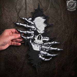 May include: An embroidered patch featuring a white skull emerging from a black, jagged-edged background, framed by two skeletal hands with long, pointed fingers reaching inward from the sides, rendered in high-contrast white thread on black fabric.