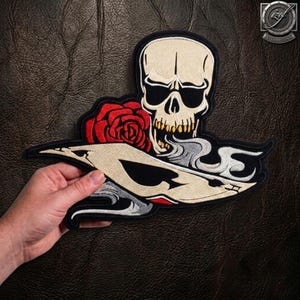 May include: An embroidered patch featuring a beige skull with dark eye sockets and teeth, positioned above a red rose and a beige spade symbol. The design includes swirling grey smoke accents and a black border on a dark textured background.