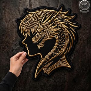 Puede incluir: Un parche bordado negro con un diseño de hilo dorado de una cabeza de dragón integrada en una silueta de perfil humano. El dragón presenta patrones geométricos complejos y escamas, con un borde negro definido.