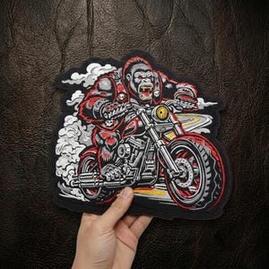 May include: Embroidered patch featuring a gorilla riding a red motorcycle. The gorilla is depicted with red fur and a fierce expression, wearing a skull-adorned vest. The motorcycle has a yellow headlight and a winding road graphic. The patch is black with white and red accents.