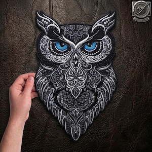 May include: An embroidered owl patch featuring intricate silver and white filigree patterns on a black background. The owl has striking, vivid blue eyes. The design is detailed with swirling lines and geometric textures, creating a bold, high-contrast appearance suitable for fabric application.