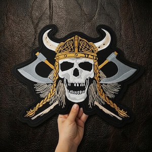 May include: Embroidered patch featuring a Viking skull wearing a horned helmet and crossed axes. The skull is white with a detailed design, and the helmet is gold and black. The axes are silver and gold, set against a black background.