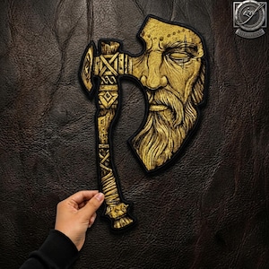 May include: A gold-colored embroidered patch featuring a detailed axe head design integrated with a bearded face. The axe handle is wrapped in textured patterns, and the entire piece is outlined with a black border against a dark background.