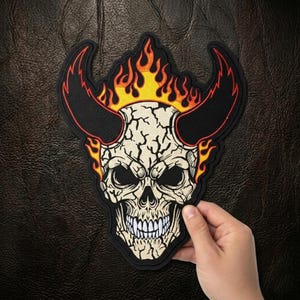 May include: A black, white, and red skull patch with flames and horns. The skull has a cracked appearance and a menacing expression. The patch is held by a hand against a dark leather background.
