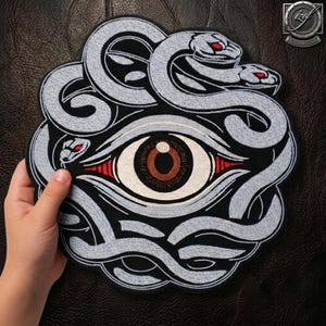 May include: A circular embroidered patch featuring a central human eye with a brown iris and black pupil, surrounded by a coiled light grey snake with red eyes. The design is set against a black background with intricate stitching details.