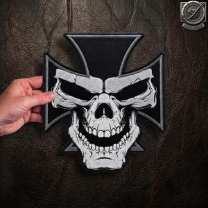 Peut inclure: Un écusson brodé noir et blanc représentant un crâne stylisé centré sur une forme de croix. Le crâne présente des orbites profondes et une mâchoire large et souriante. Le motif est réalisé avec un fil contrasté et des ombrages détaillés.