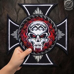 Puede incluir: Un parche bordado en blanco y negro con forma de cruz y un emblema circular central. El emblema presenta una calavera blanca con ojos rojos y colmillos, rodeada por un patrón de remolinos rojos y negros sobre un fondo negro.