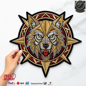 Könnte beinhalten: Ein kreisförmiger bestickter Aufnäher mit einem stilisierten Wolfskopf mit geometrischen Details aus goldenen, silbernen und roten Fäden. Der Wolf ist mit offenem Maul und sichtbaren Zähnen dargestellt, eingefasst in einen sternförmigen Rahmen.