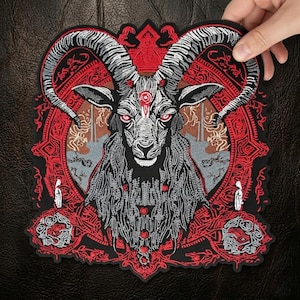 May include: Embroidered patch featuring a detailed illustration of a goat's head with large, curved horns. The design incorporates red, gray, and black colors with intricate patterns and a central eye symbol. The patch is held by a hand.