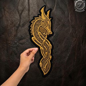 Peut inclure: Un écusson brodé représentant un dragon de couleur dorée avec des motifs de nœuds complexes sur un fond noir. Le dragon est représenté sous une forme stylisée et allongée avec des lignes courbes et des textures détaillées sur tout le corps.
